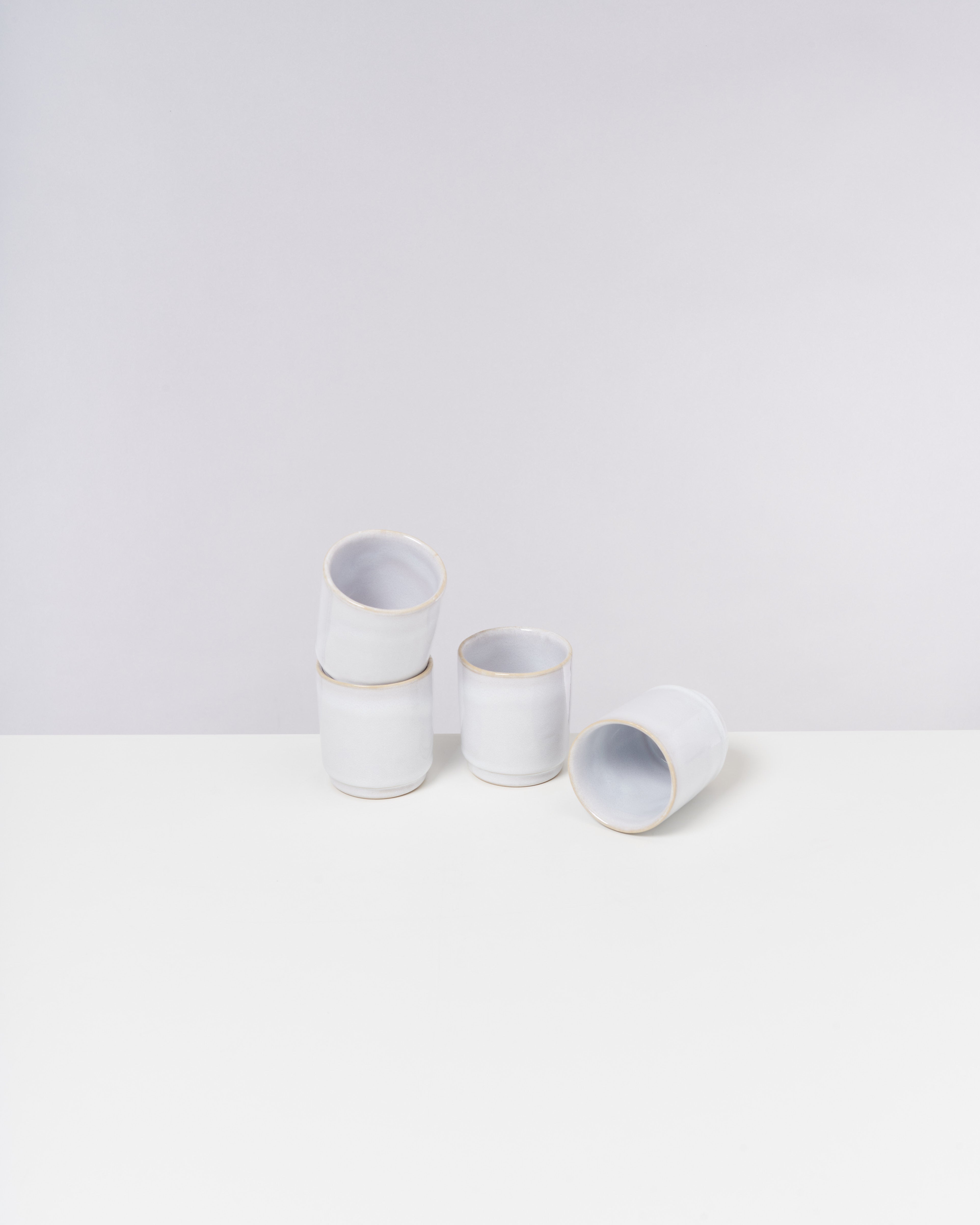 Fünf weiße Becher aus Keramik stehen und liegen auf einem weißen Tisch vor einem grauen Hintergrund.