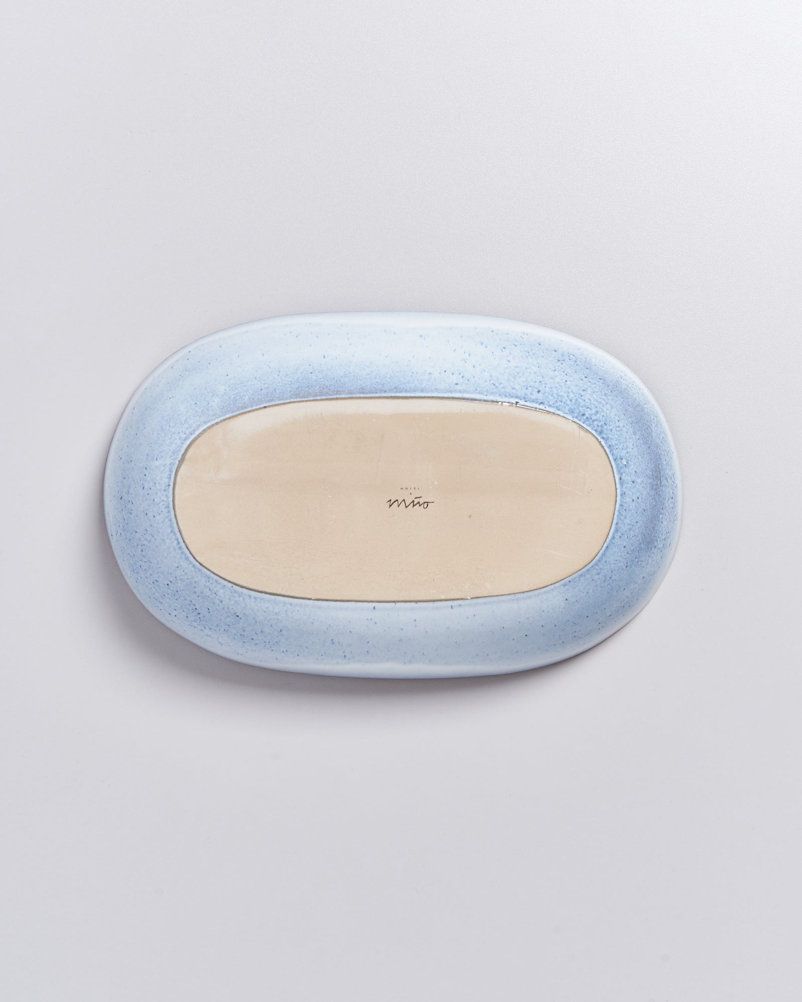 Ovale, flache Keramikplatte mit hellem Zentrum und bauchigem hellblauen Rand auf weißem Hintergrund.