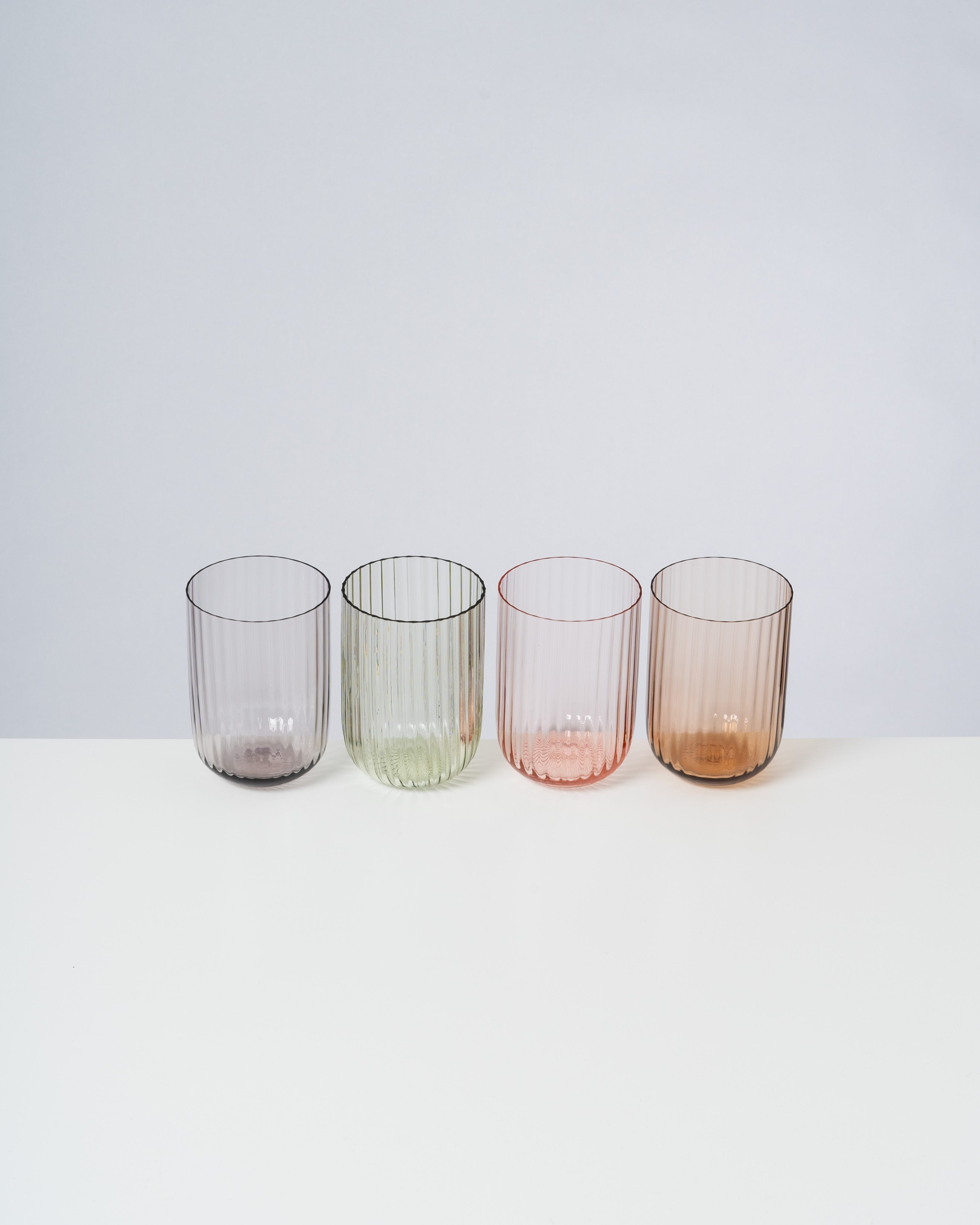 Vier verschiedenfarbige geriffelte Gläser stehen nebeneinander auf einem weißen Tisch.