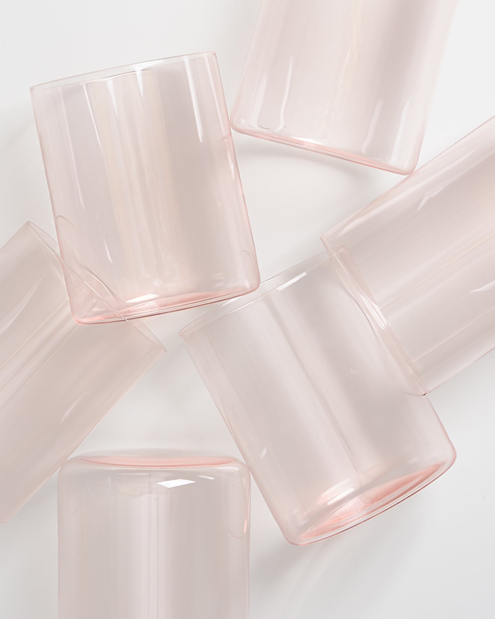 Sechs rosafarbene Gläser liegen auf einer weißen Fläche, schräg zueinander platziert.