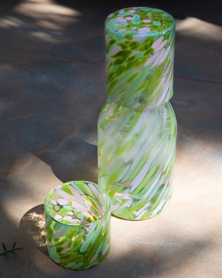 Karaffe mit Glas in Grün- und Rosatönen auf Schatten-und-Licht-Musterung.