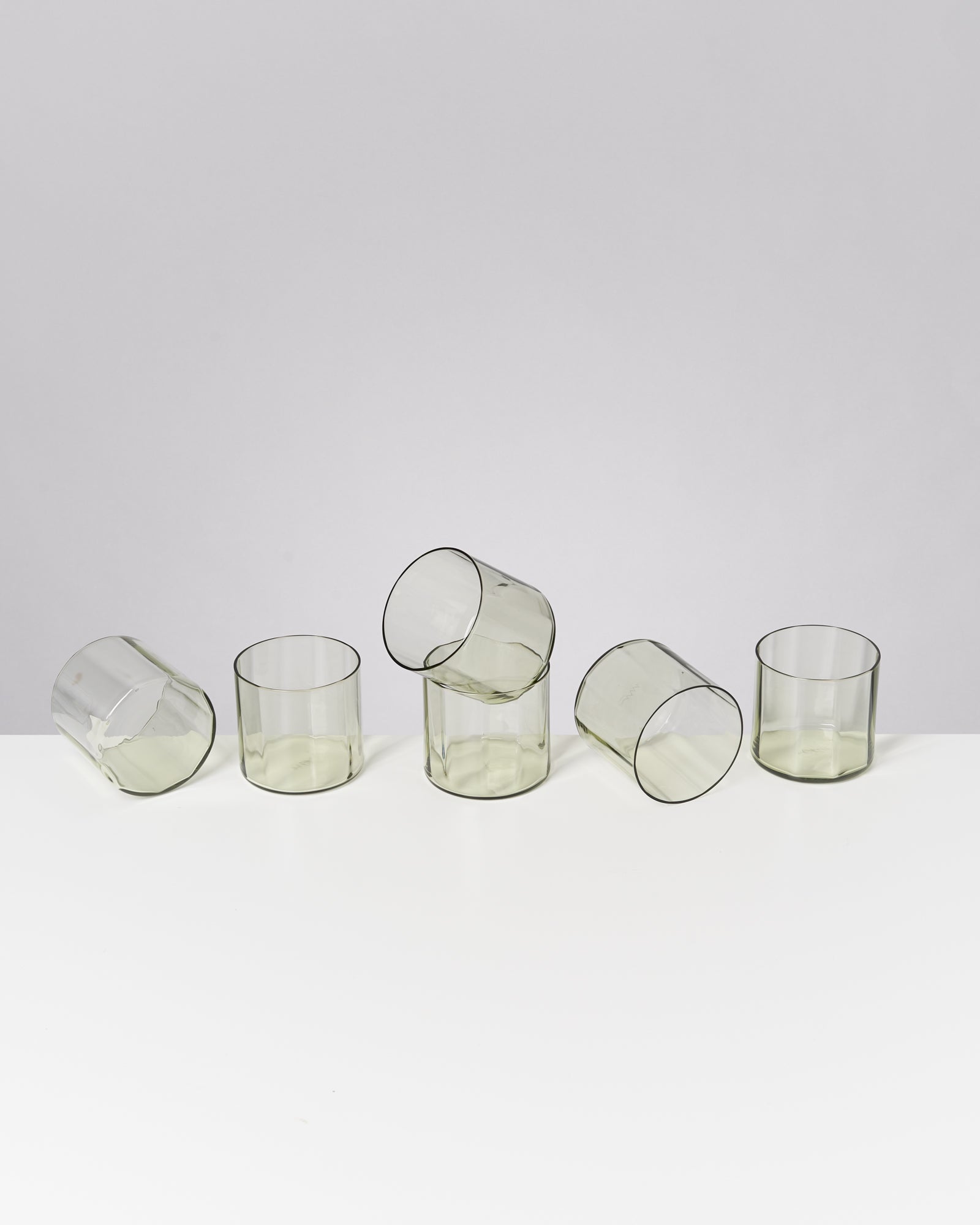 Fünf transparente Gläser in verschiedenen Positionen auf einem weißen Tisch vor grauem Hintergrund.