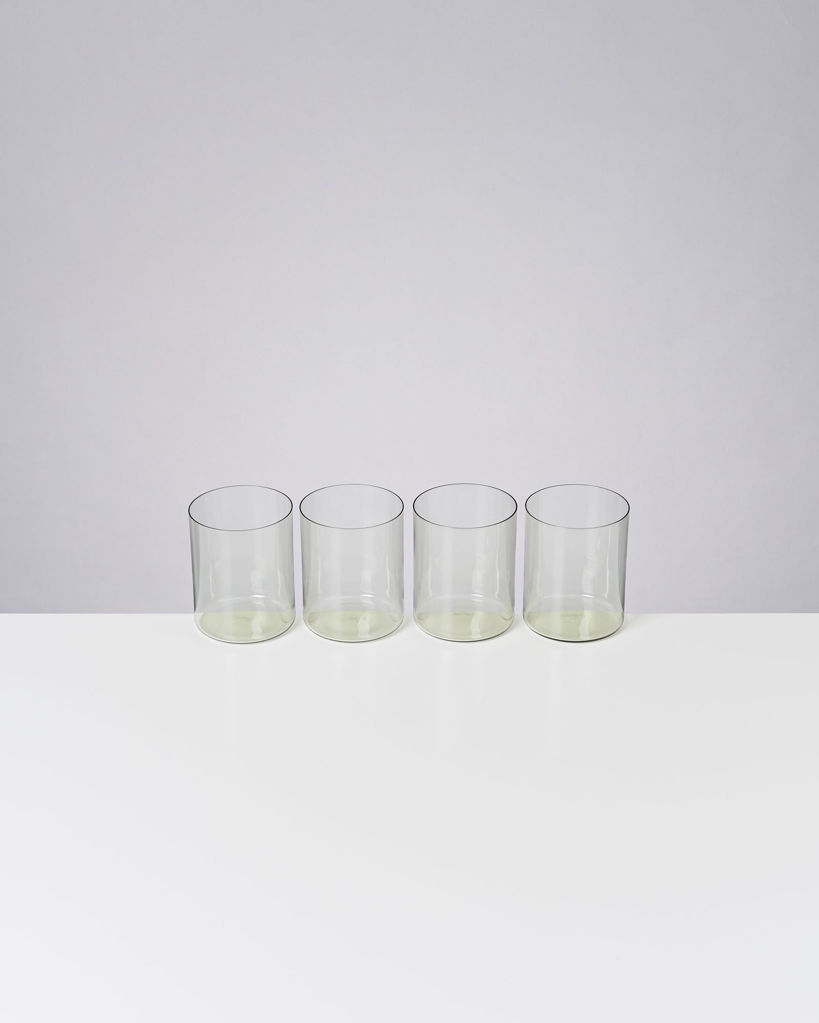 Vier leere, klare Gläser stehen nebeneinander auf einem weißen Tisch vor einem grauen Hintergrund.