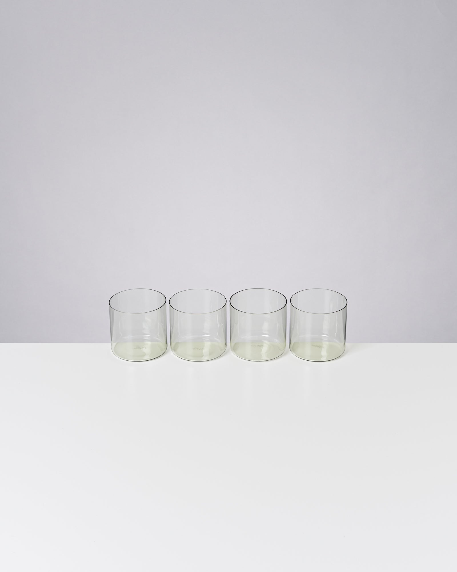 Vier leere, klare Trinkgläser stehen nebeneinander auf einer weißen Oberfläche vor grauem Hintergrund.