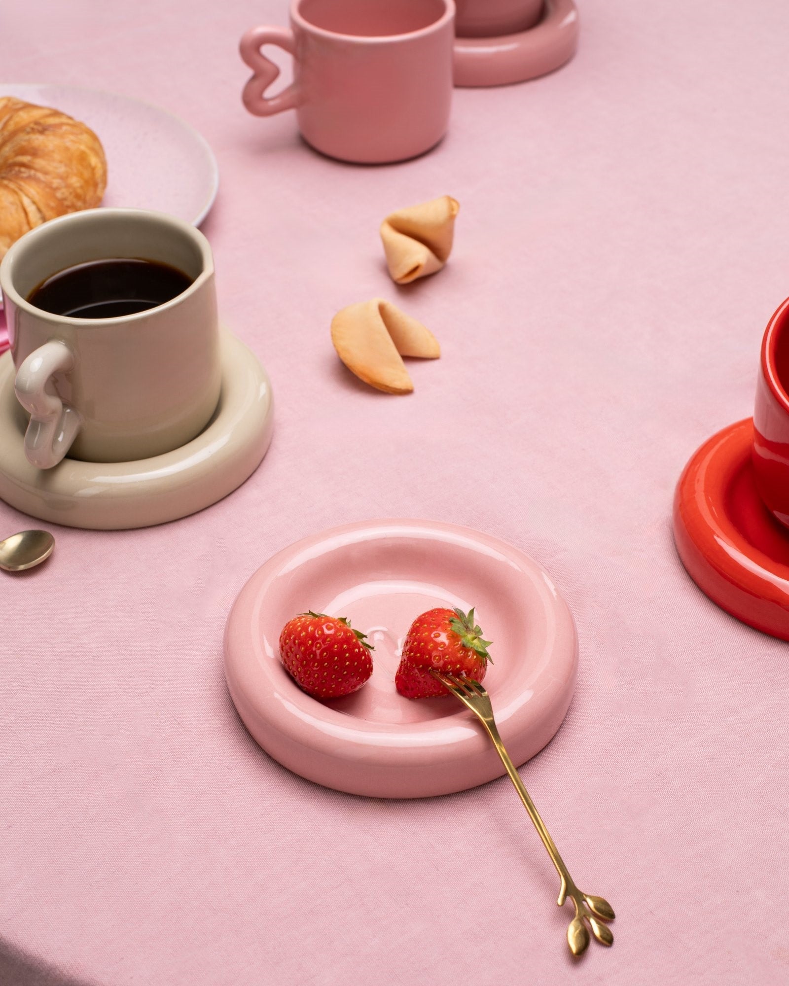 Auf einem rosa Tischtuch stehen Tassen, Croissants, Glückskekse und ein Teller mit Erdbeeren neben einer goldenen Gabel.