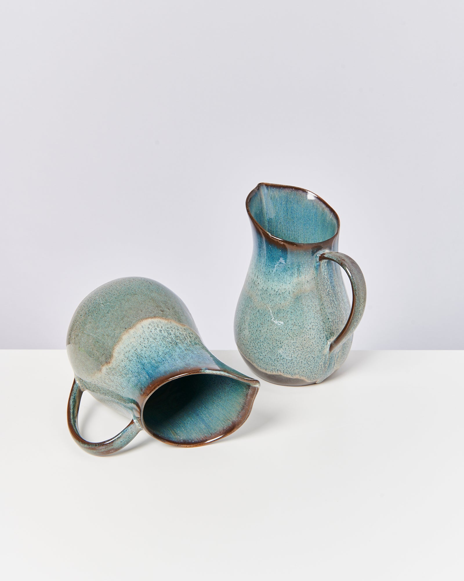 Zwei blau-grüne Keramikkrüge, einer stehend und einer liegend, auf einer weißen Oberfläche.