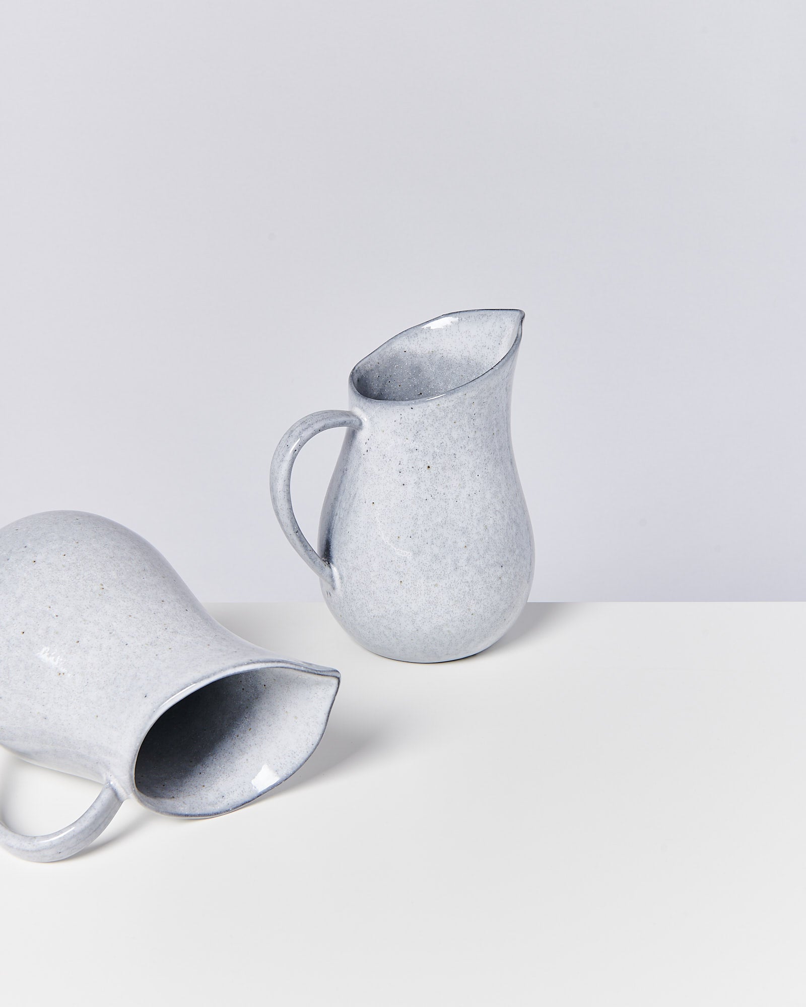Zwei graue Keramikkrüge, einer stehend und einer liegend, auf einer weißen Fläche vor grauem Hintergrund.