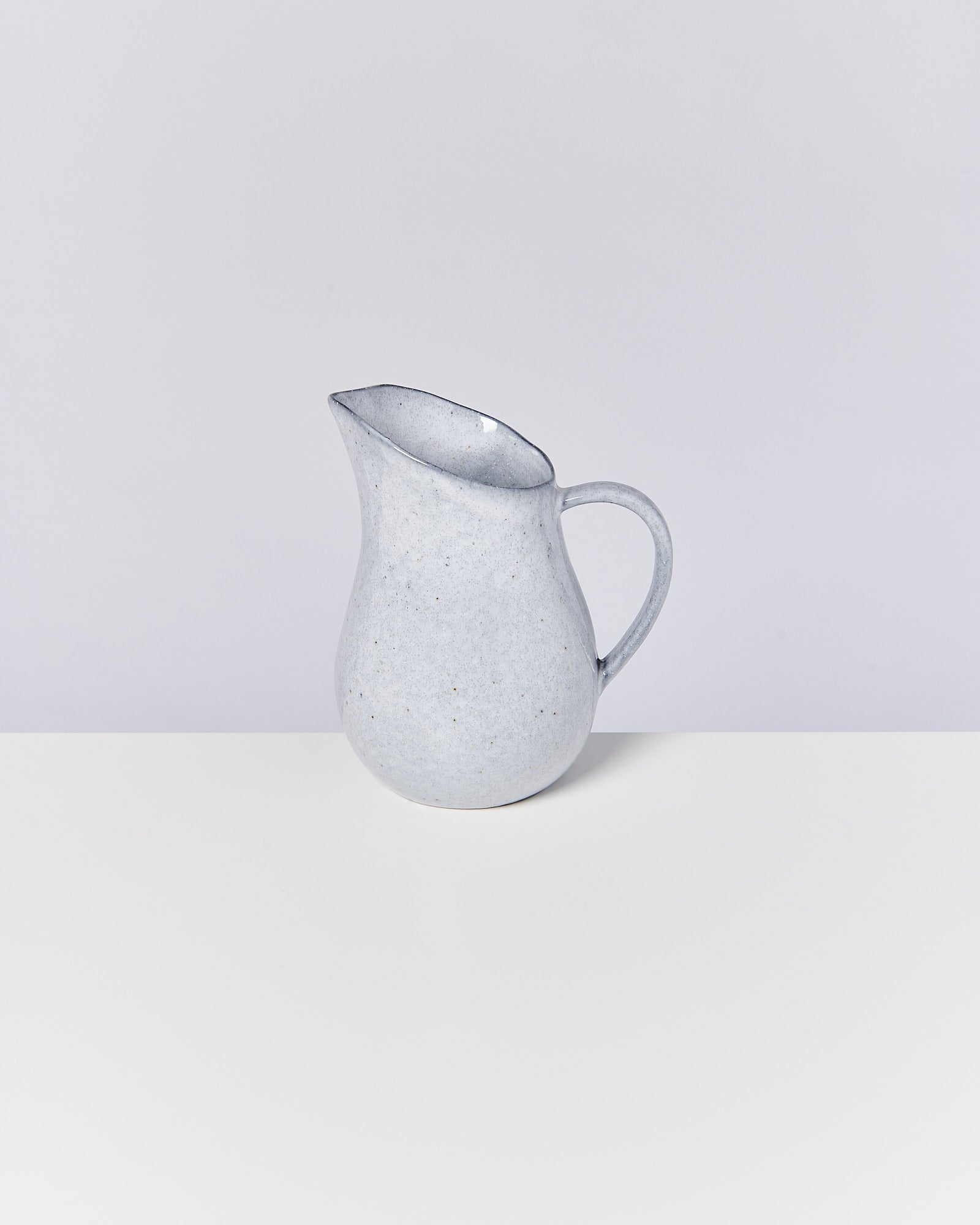 Grauer Keramikkrug mit Henkel auf weißem Tisch vor einem neutralen Hintergrund.