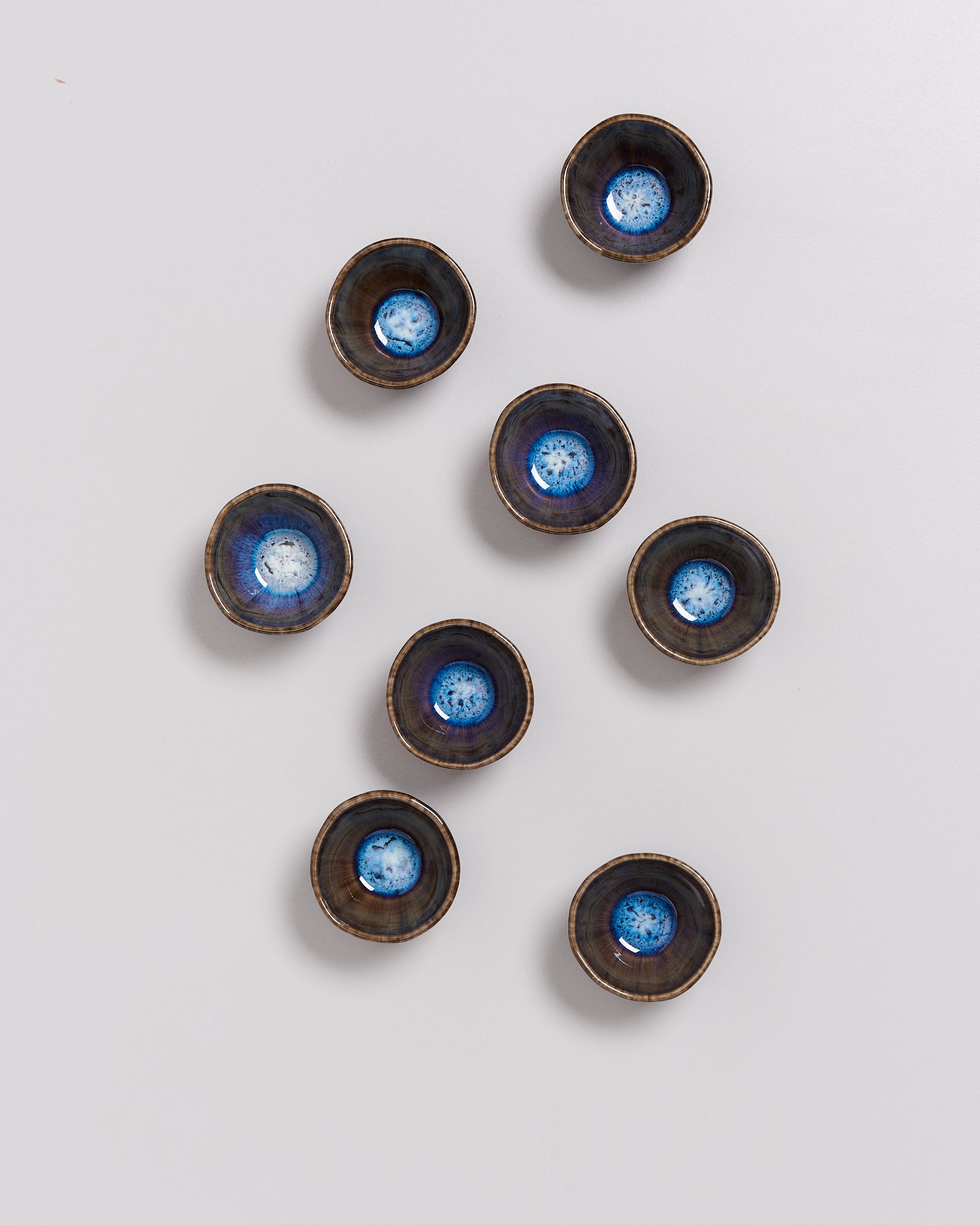 Acht kleine keramische Schalen mit blauem und braunem Muster sind auf einer weißen Fläche angeordnet.