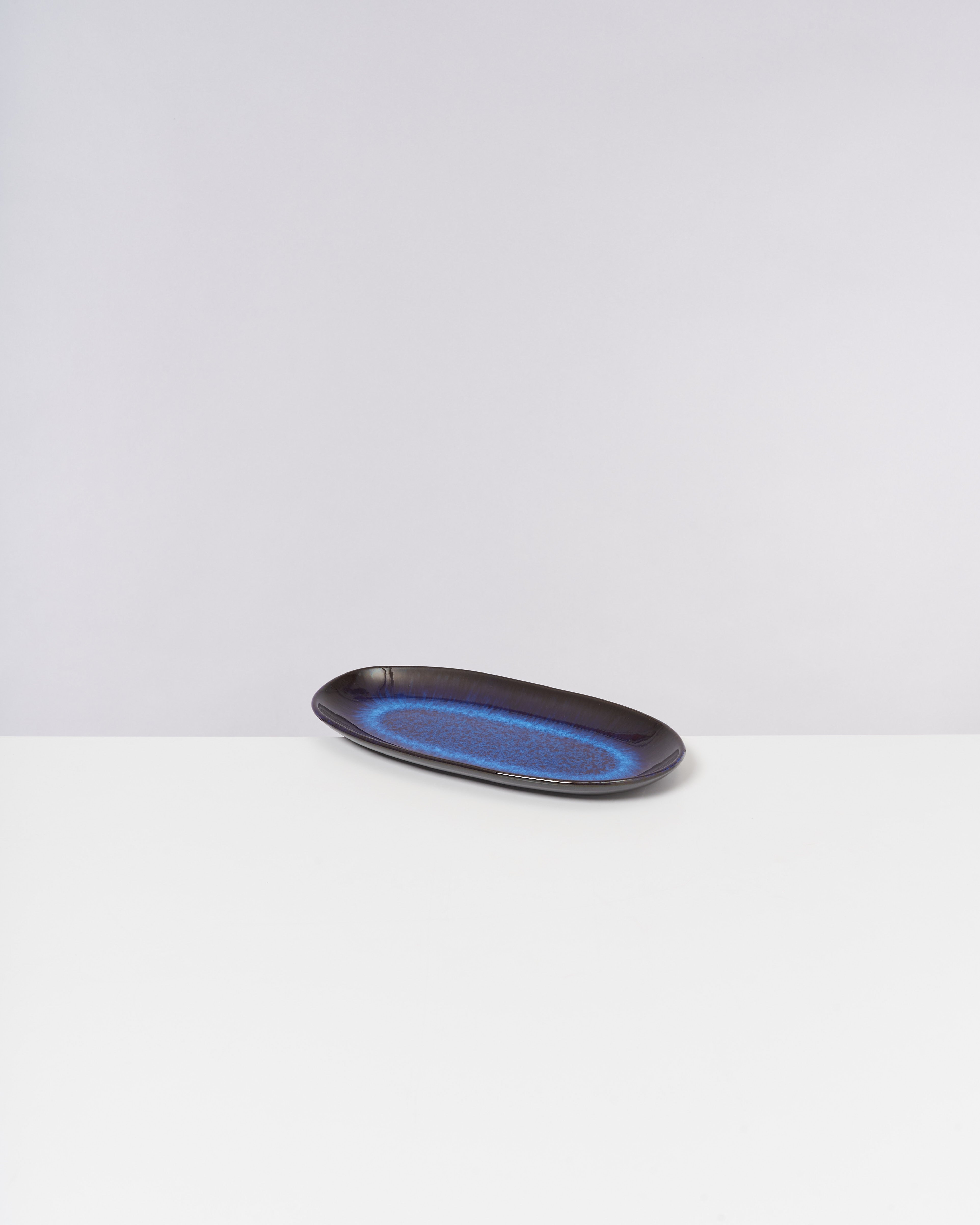 Ein länglicher, schwarz-blauer Keramikteller liegt auf einer weißen Oberfläche vor einem grauen Hintergrund.