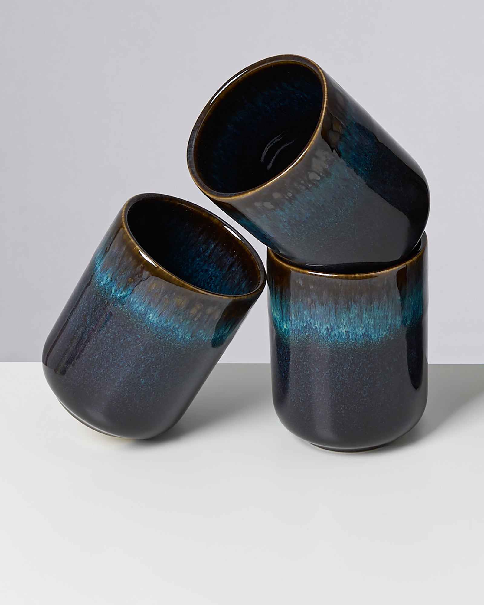 Drei dunkelblaue Keramikbecher mit türkisfarbenem Verlauf stehen auf einer weißen Oberfläche vor grauem Hintergrund.