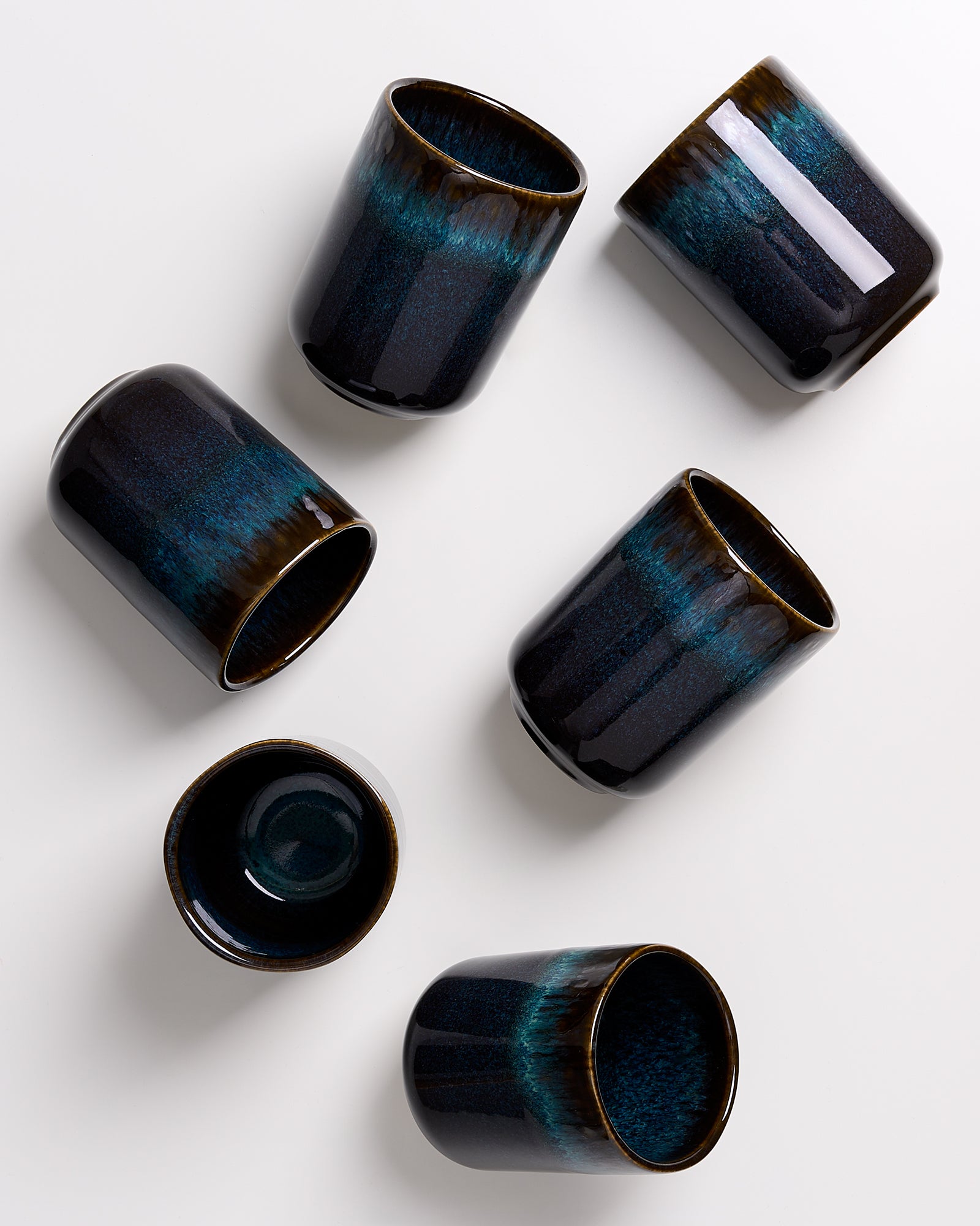 Sechs schwarze Keramikbecher mit blauer Glasur auf weißem Hintergrund verstreut.