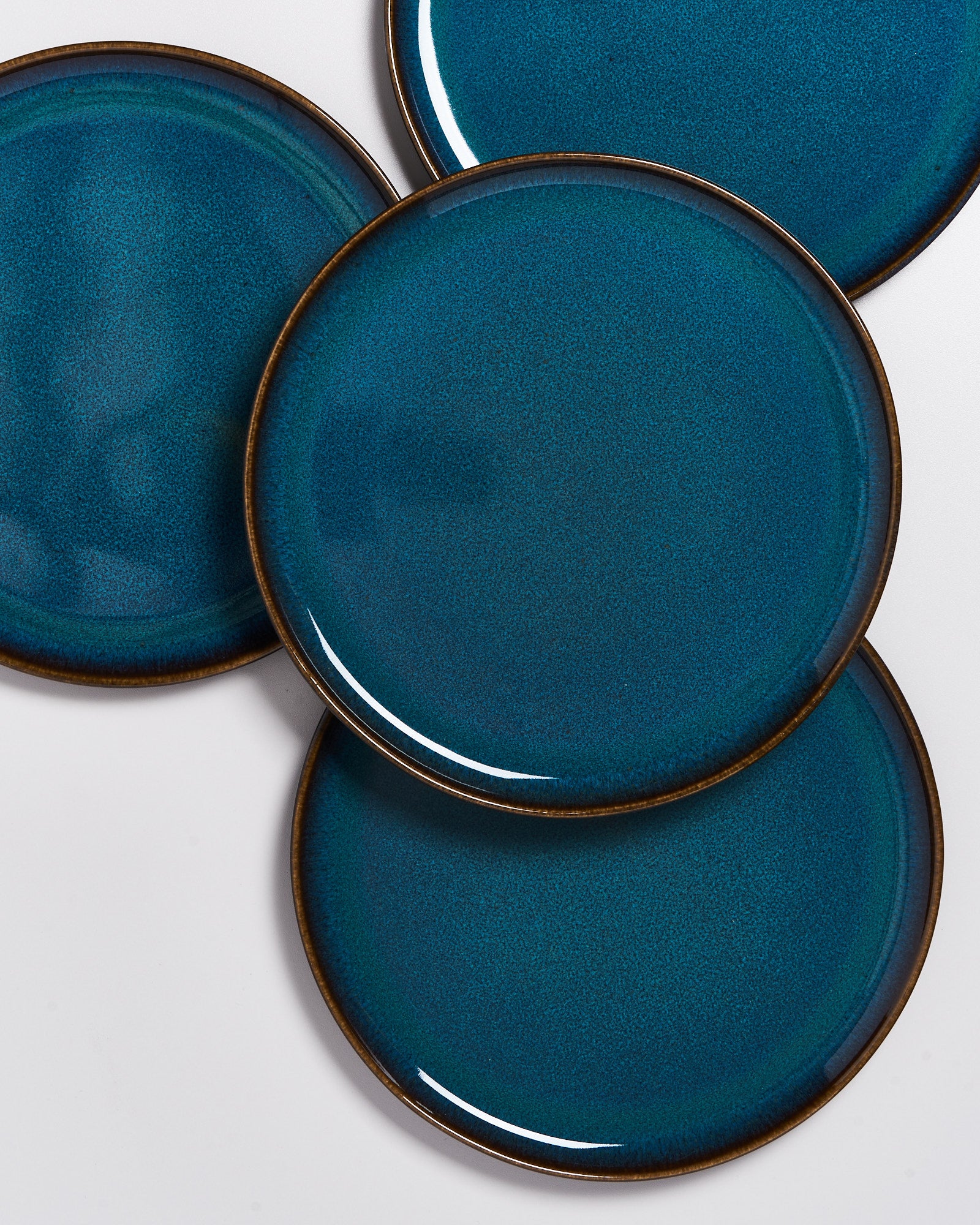 Vier übereinanderliegende, runde, blau-grüne Teller mit braunem Rand auf weißem Hintergrund.