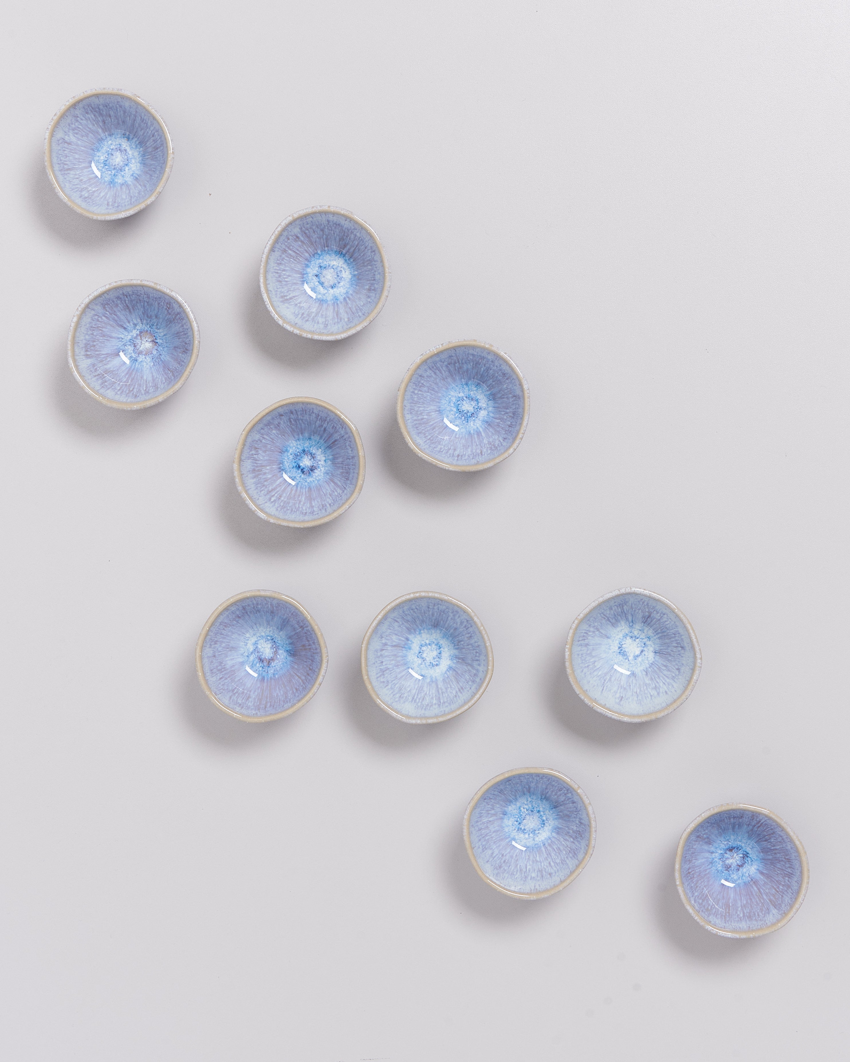 Zehn kleine, blaue Keramikschalen sind in einem diagonalen Muster auf einem hellgrauen Hintergrund angeordnet.