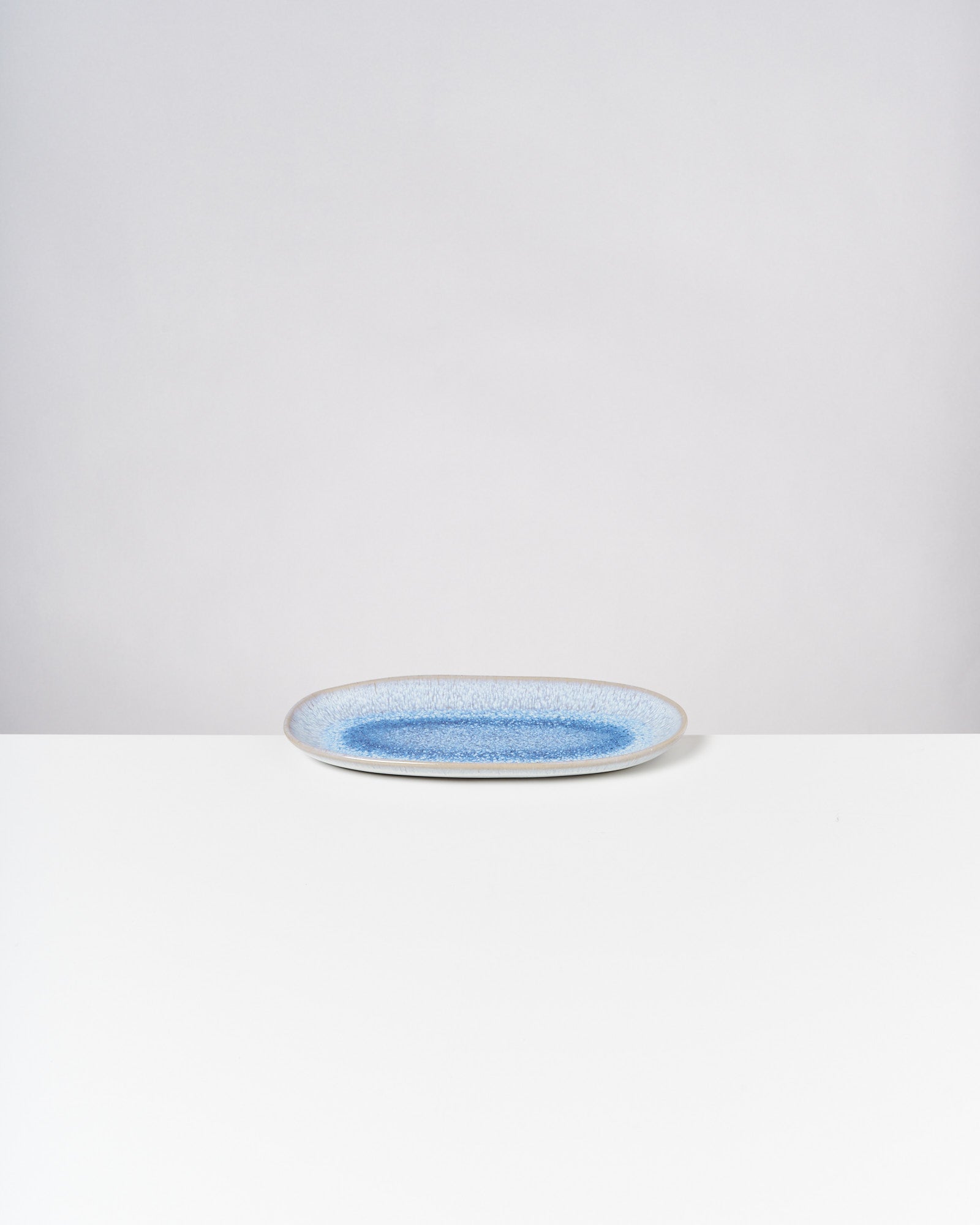 Ovale Keramikplatte mit blauer Mitte auf weißem Tisch vor grauem Hintergrund.