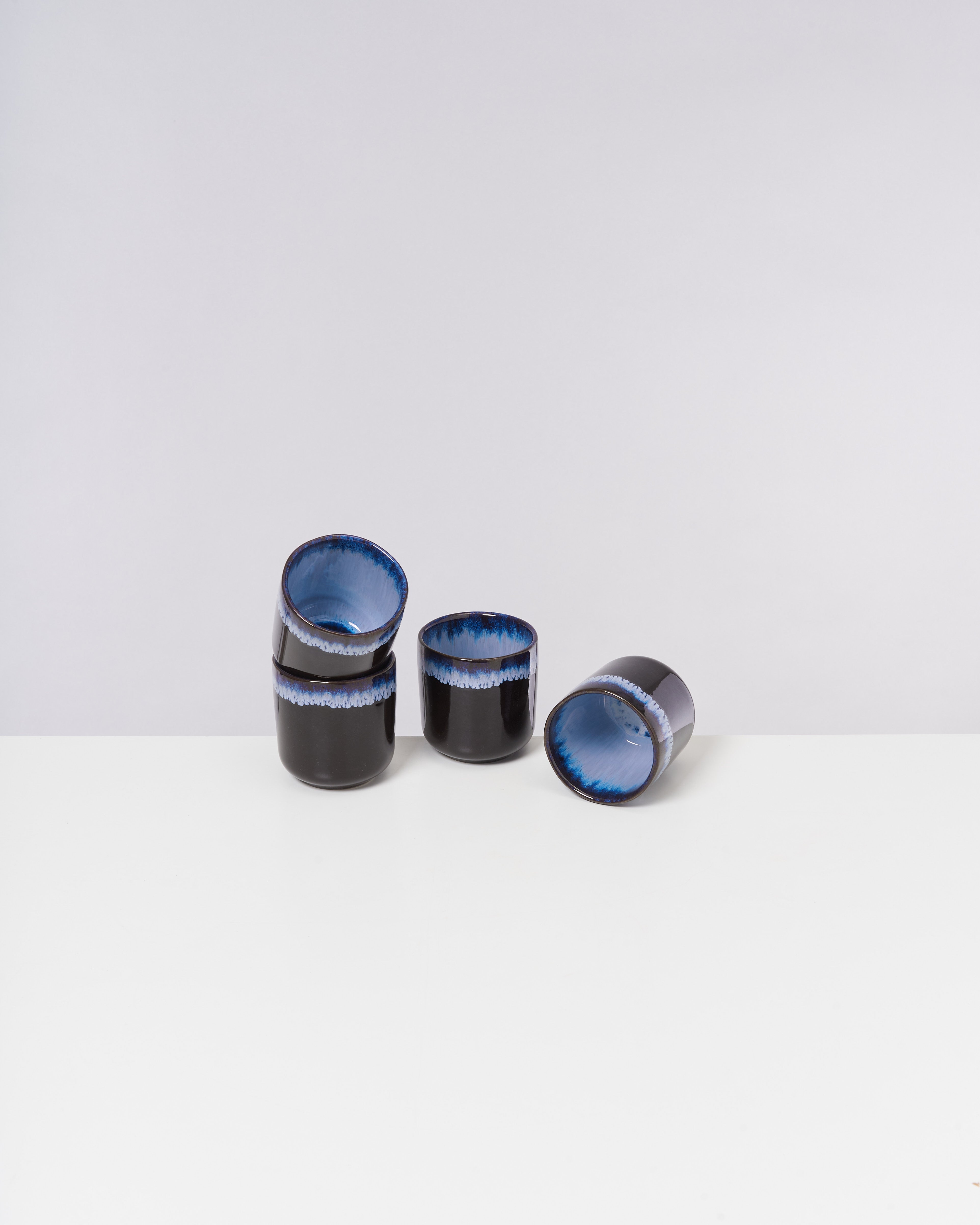 Fünf schwarze Keramikbecher mit blauer Glasur innen und am Rand, auf einem weißen Tisch angeordnet.