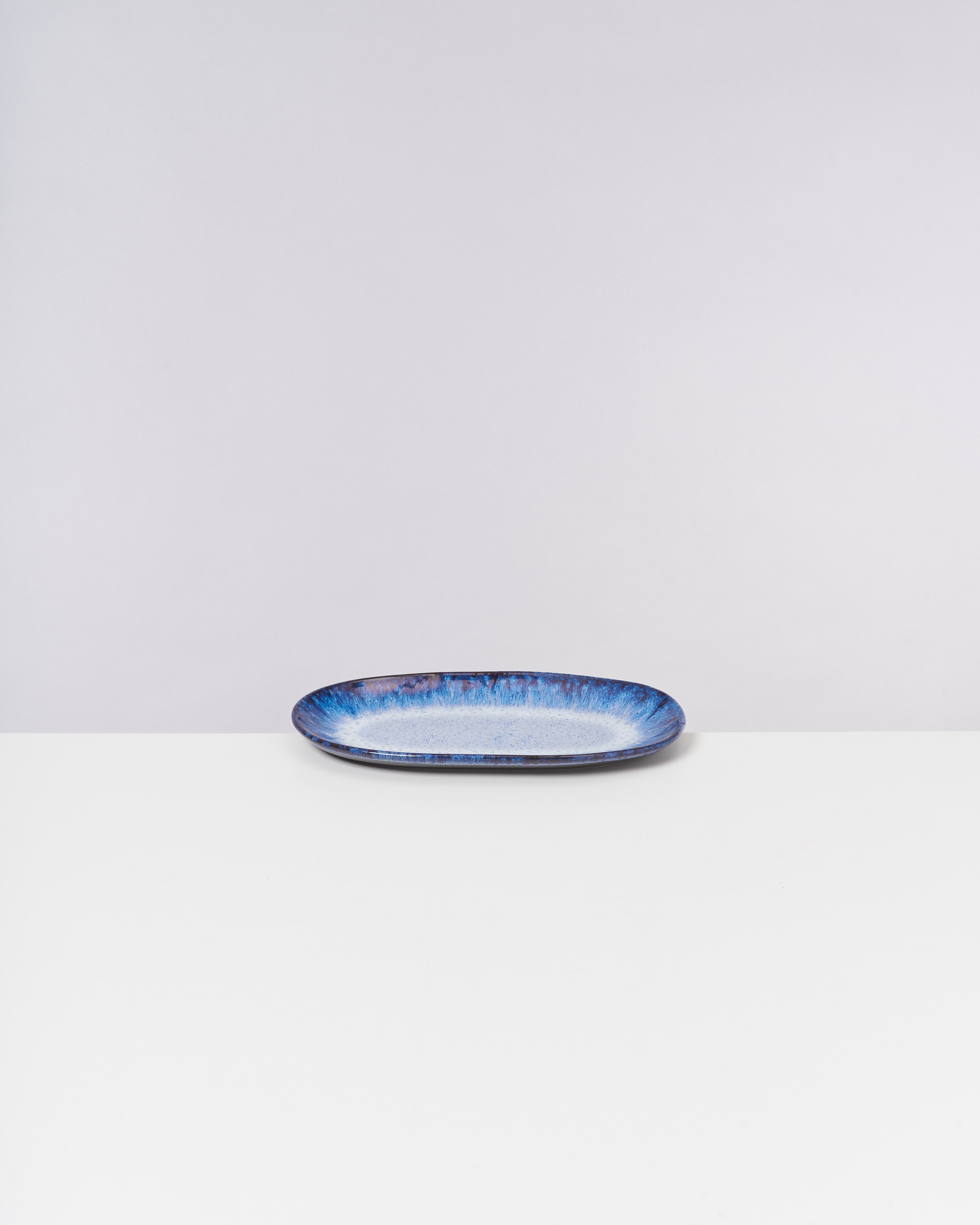 Eine ovale, blau-weiße Keramikplatte auf einem weißen Tisch vor grauem Hintergrund.