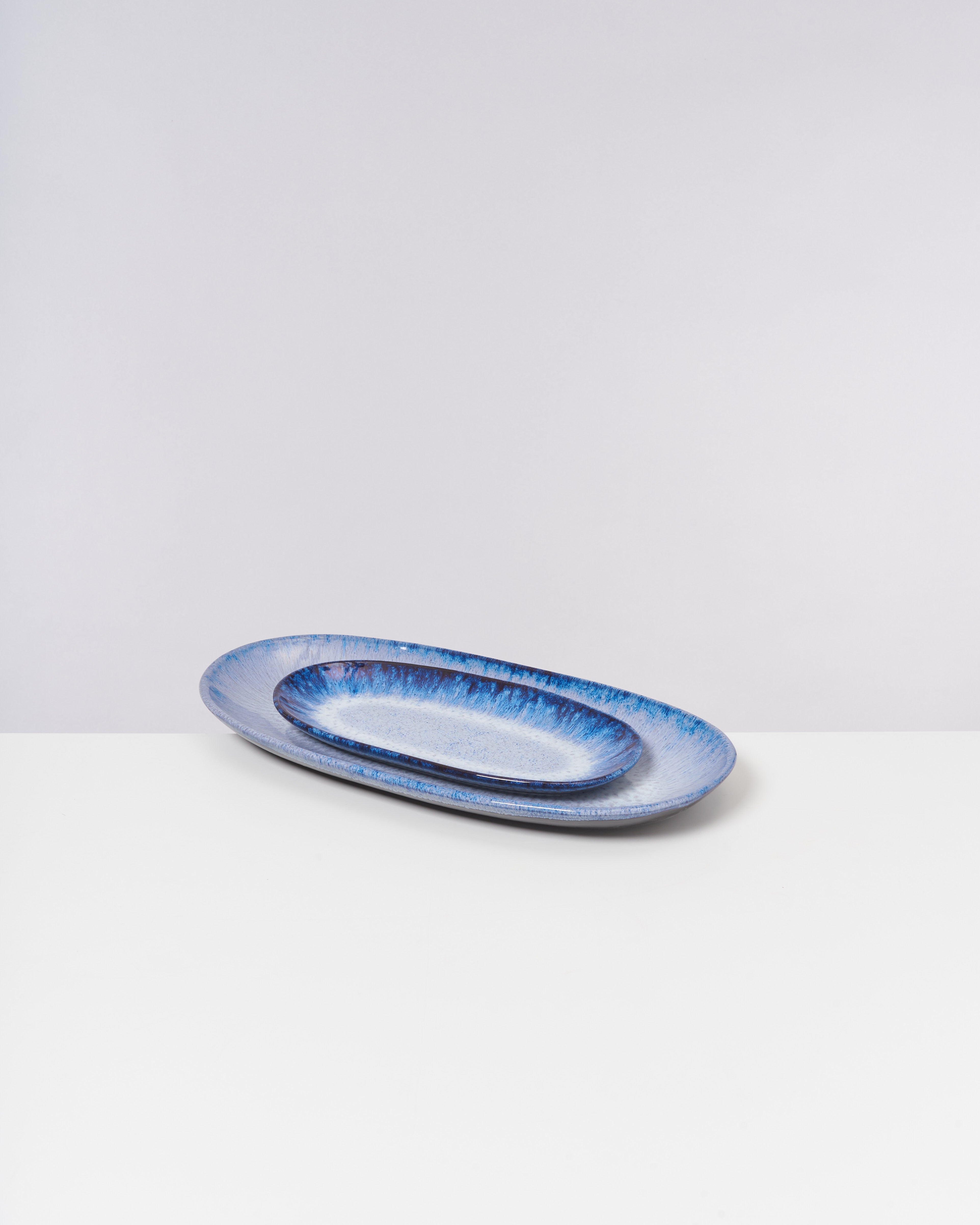 Zwei ovale, blaue Keramikteller liegen ineinander auf einer weißen Oberfläche vor einem hellen Hintergrund.
