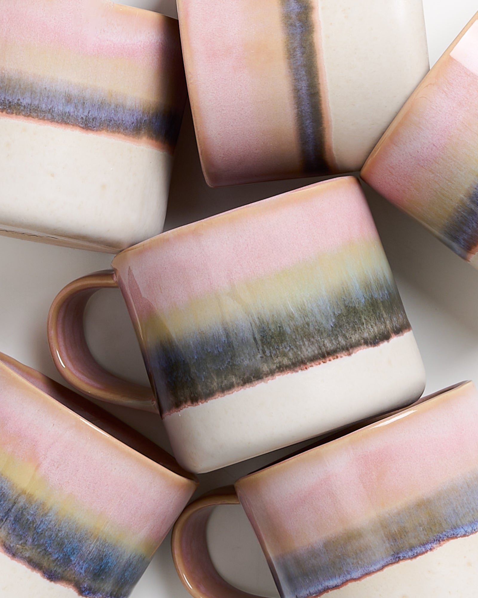 Mehrere Keramiktassen mit pastellfarbenen Streifen in Rosa, Beige und Blau liegen übereinander auf weißem Hintergrund.