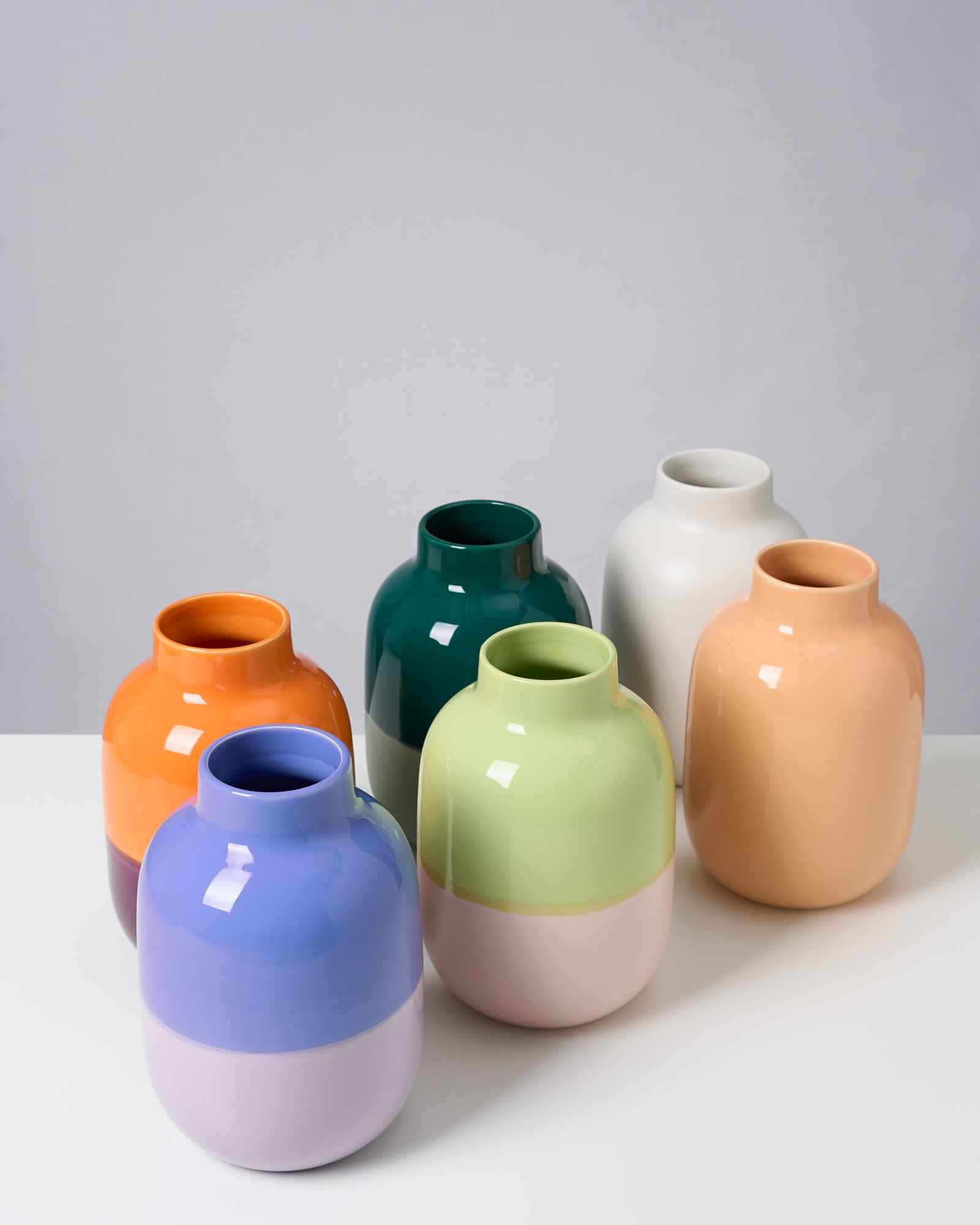 Sechs bunte, glänzende Vasen stehen auf einem weißen Tisch vor einem grauen Hintergrund.