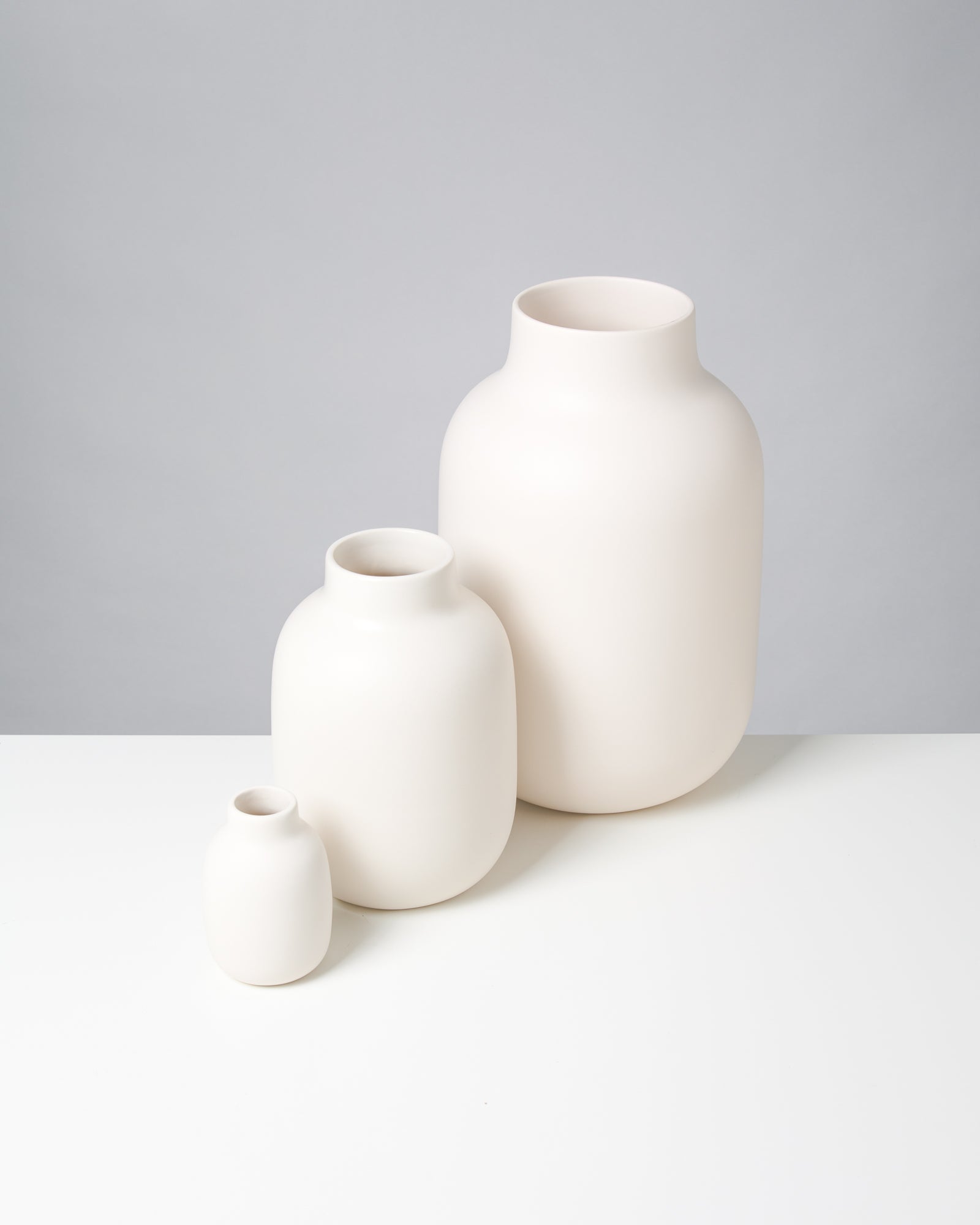 Drei weiße Vasen unterschiedlicher Größe stehen auf einem weißen Tisch vor einem grauen Hintergrund.