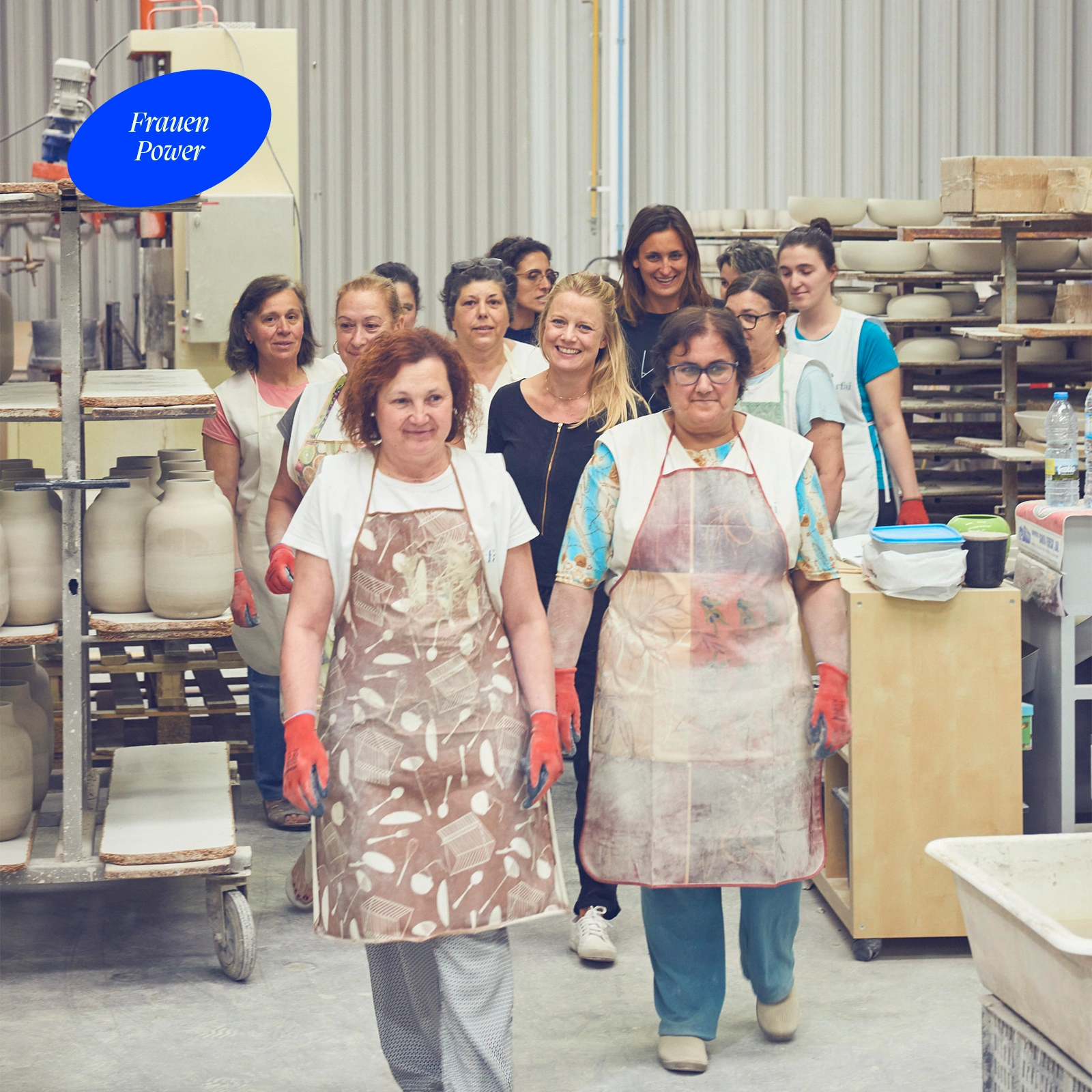Eine Gruppe von Frauen in einer Töpferwerkstatt, trägt Schürzen und lächelt. Ein Schild mit "Frauen Power" ist sichtbar.