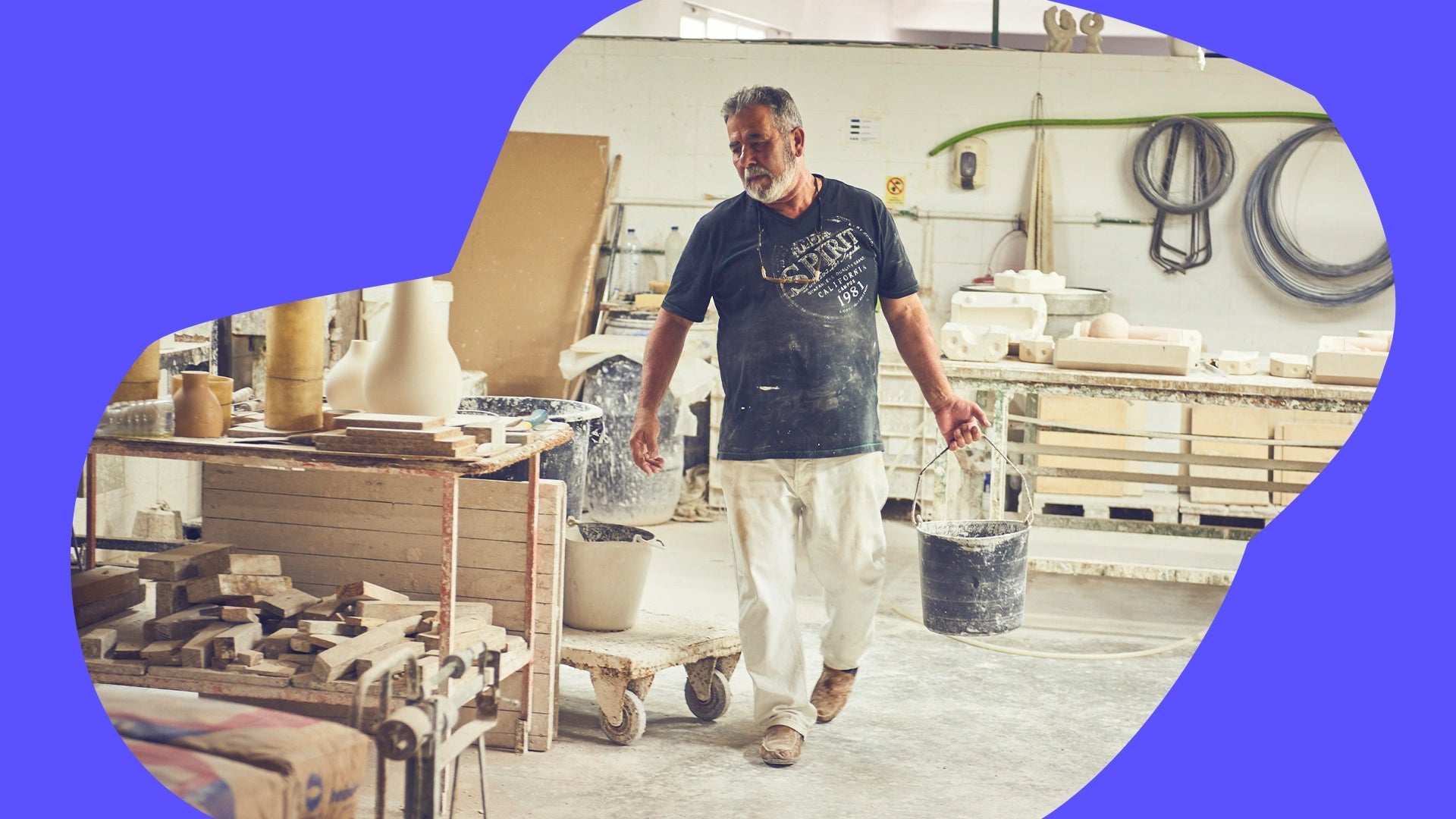 Ein Mann in einer Werkstatt trägt einen Eimer, umgeben von Keramikformen und Werkzeugen.