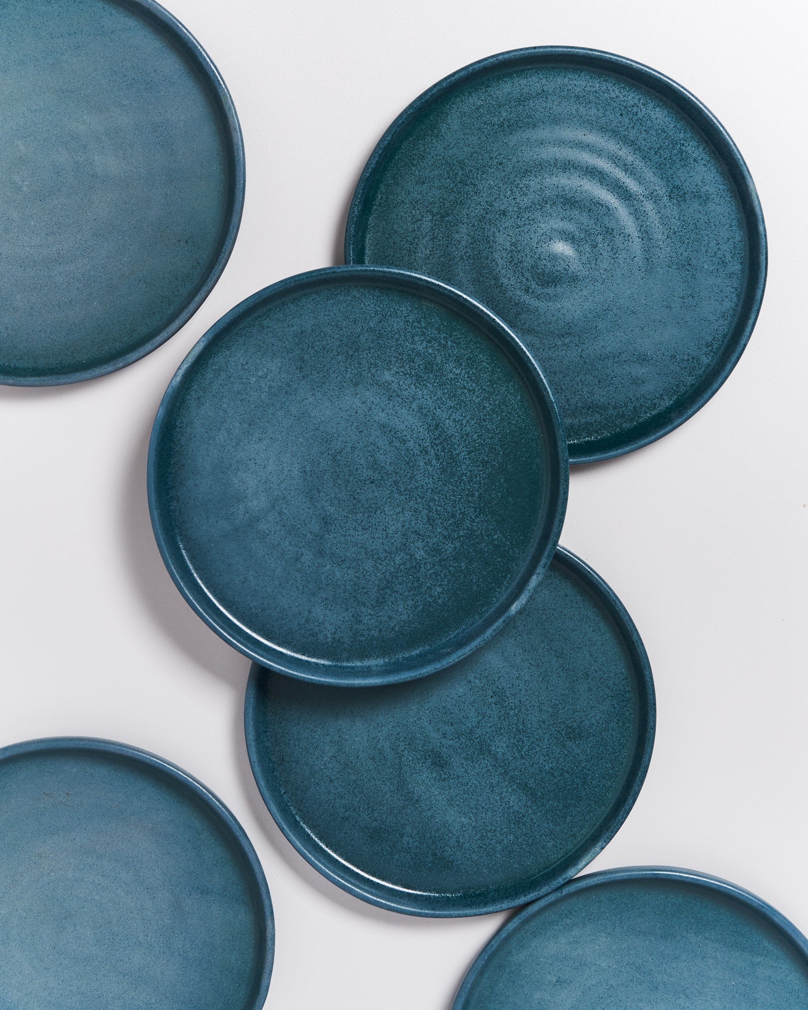 Sechs dunkelblaue Keramikteller liegen auf einem weißen Hintergrund verteilt.