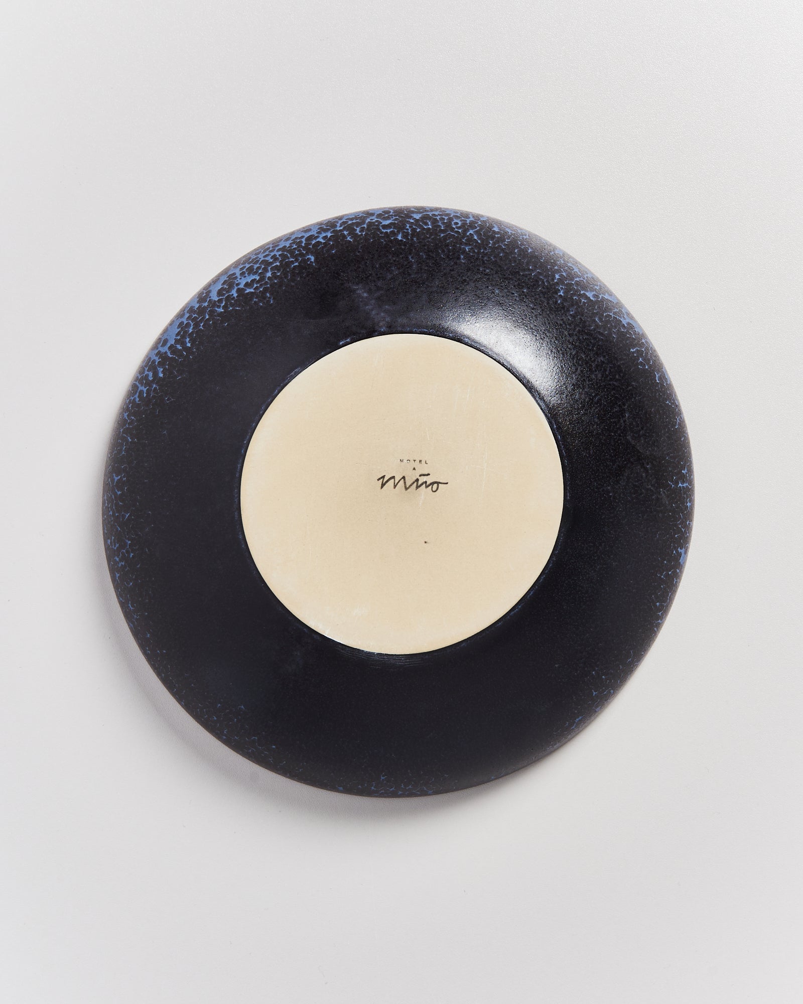 Umgedrehter Teller mit dunklem Rand und hellem Zentrum auf weißem Hintergrund; Aufschrift "Hotel Miró".