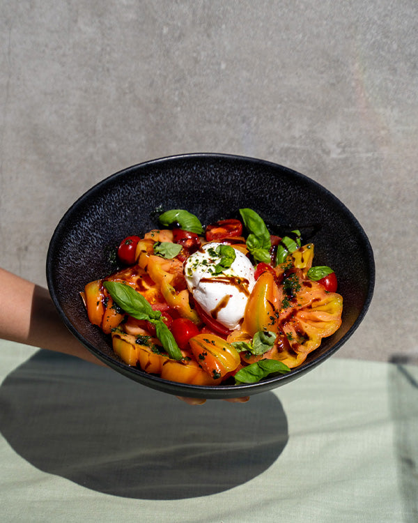 Schwarze Schale mit buntem Tomatensalat, Burrata, Basilikum und Balsamico-Glasur, gehalten von einer Hand.