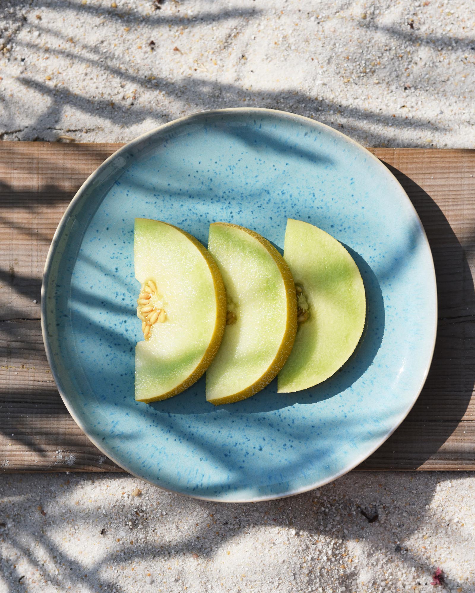 Drei Melonenscheiben auf einem blauen Teller, der auf sandigem Untergrund steht.