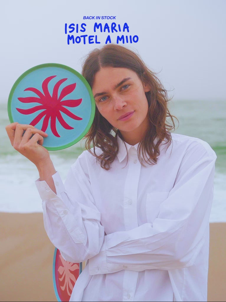 Frau in weißem Hemd hält einen bunt gemusterten Teller am Strand mit Text "ISIS MARIA MOTEL A MIIIO" darüber.
