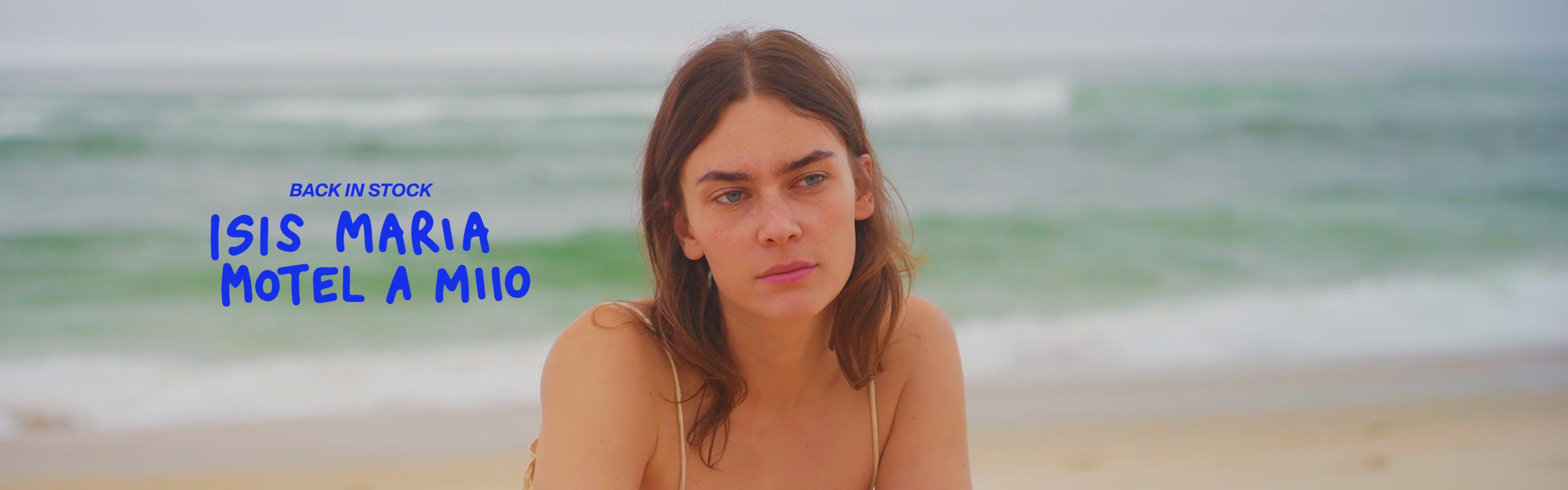 Eine Frau sitzt am Strand, im Hintergrund ist das Meer zu sehen, daneben blauer Text "Back in Stock, Isis Maria, Motel a Miio".