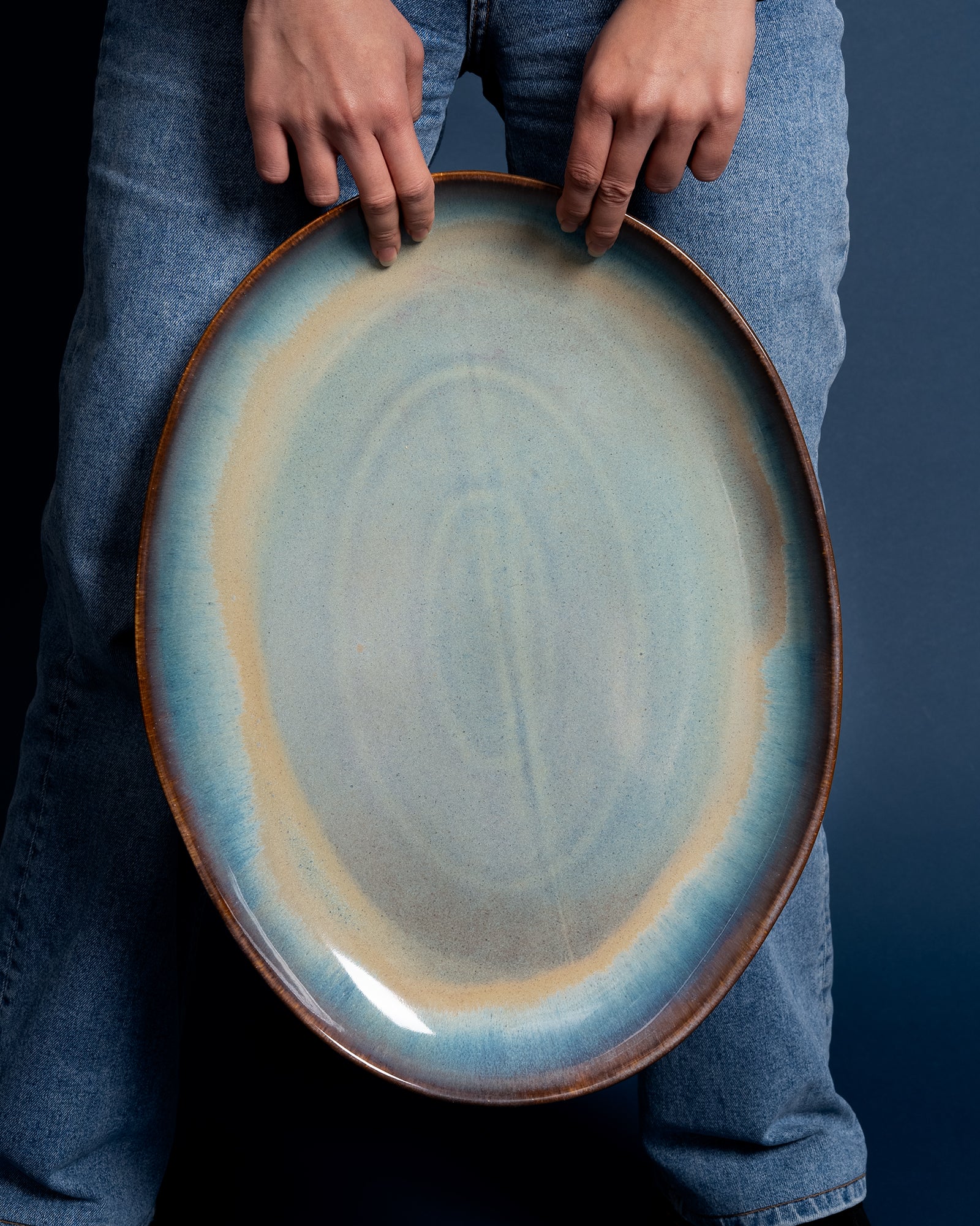 Person hält einen großen, ovalen, blau-braunen Keramikteller vor dunkelblauem Hintergrund.